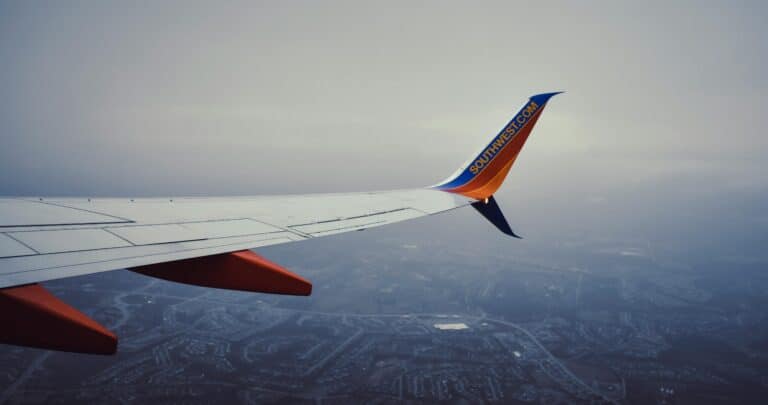 View of an airplane wing flying over a cityscape through a cloudy sky, offering a travel perspective.